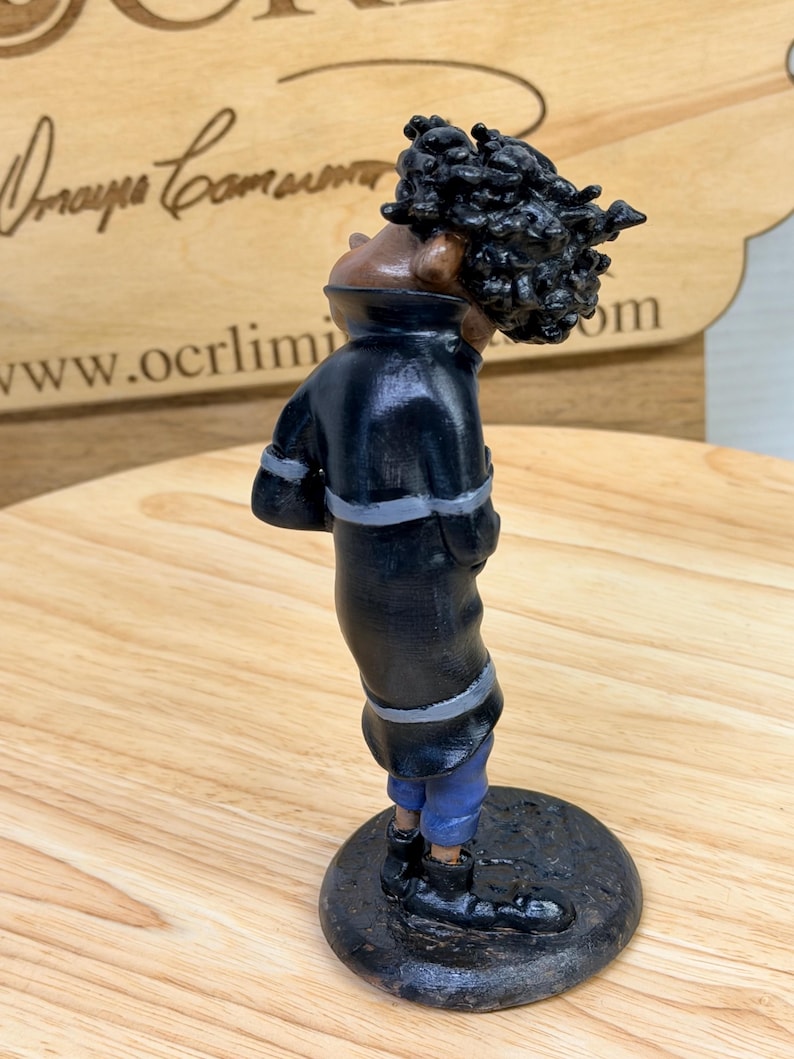 May include: A handcrafted figurine of a person in a black coat with gray trim and blue pants. The figure has a dark complexion and a large, textured hairstyle. The figurine stands on a dark, circular base.