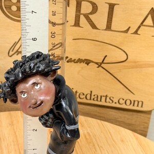 May include: A handcrafted figurine of a person wearing a black jacket and blue pants. The figurine has a detailed face with green eyes and rosy cheeks. A ruler shows the figurine is approximately 3 inches tall. The background includes the text "ORLA" and "tedarts.com".