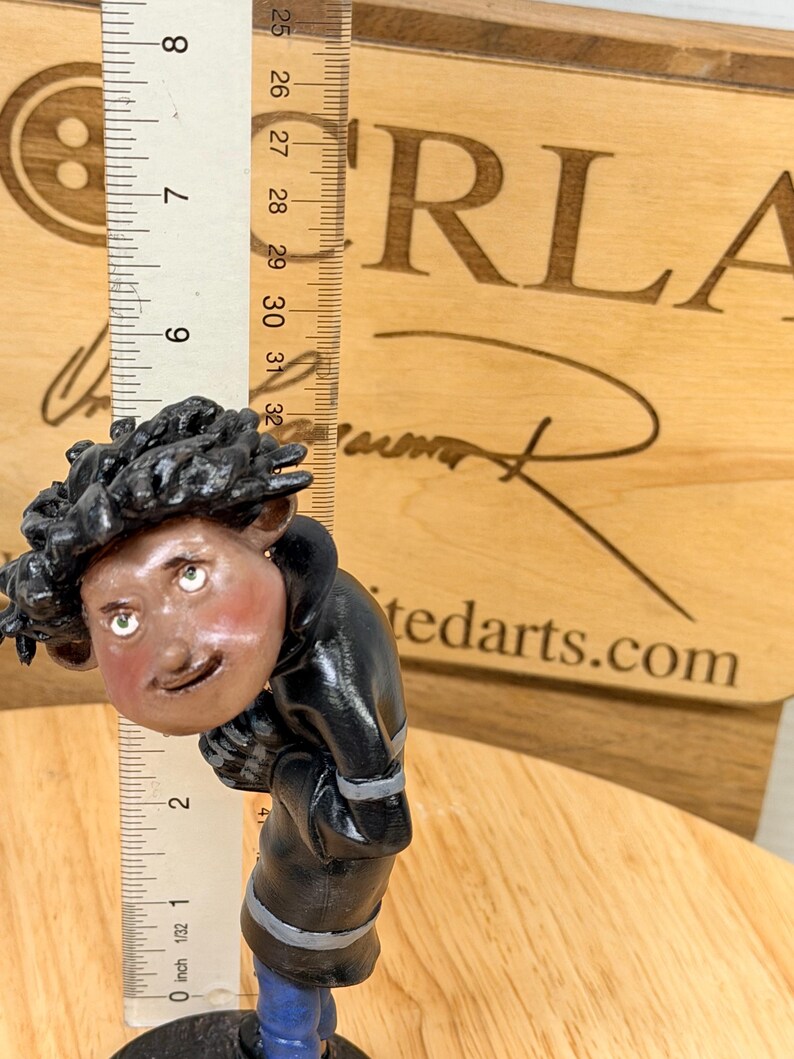 May include: A handcrafted figurine of a person wearing a black jacket and blue pants. The figurine has a detailed face with green eyes and rosy cheeks. A ruler shows the figurine is approximately 3 inches tall. The background includes the text "ORLA" and "tedarts.com".