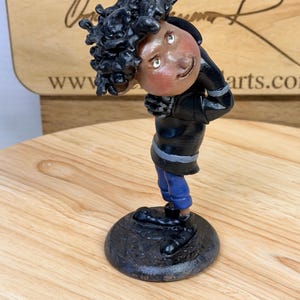 May include: A whimsical figurine of a person with dark skin and curly black hair, wearing a black jacket with silver stripes, blue pants, and black shoes. The figure is posed with a slight lean, and is mounted on a dark, textured base.