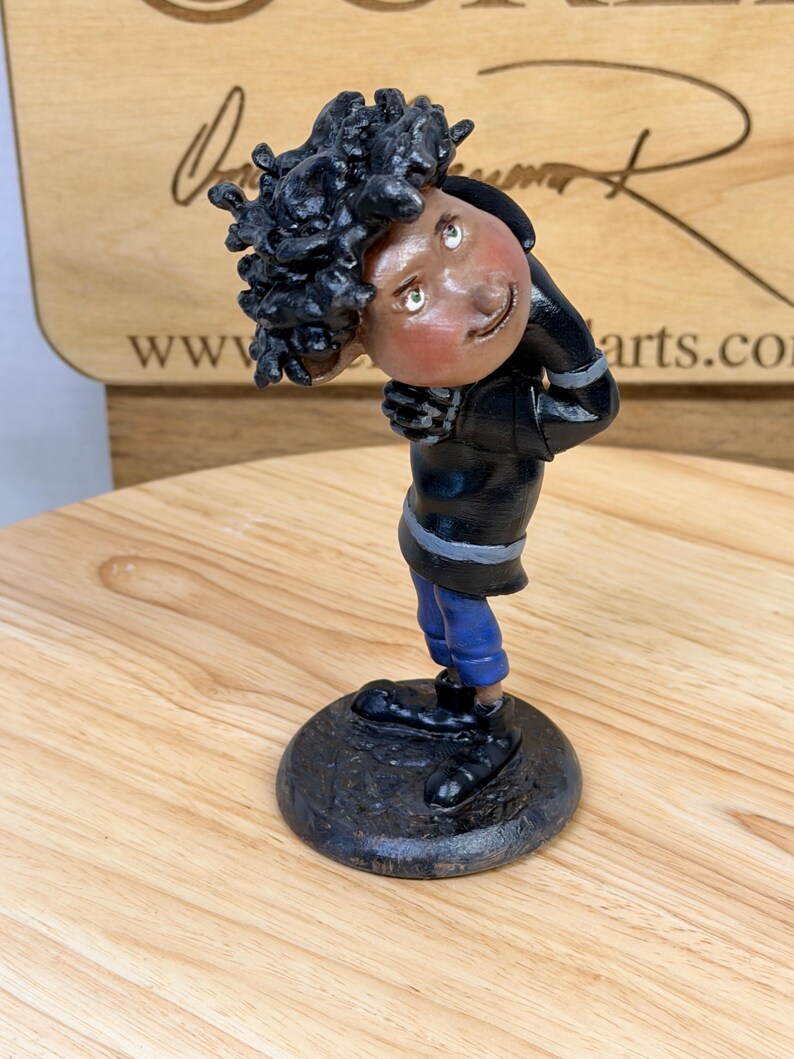 May include: A whimsical figurine of a person with dark skin and curly black hair, wearing a black jacket with silver stripes, blue pants, and black shoes. The figure is posed with a slight lean, and is mounted on a dark, textured base.