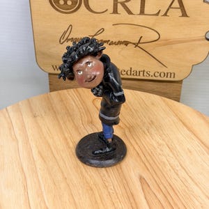 May include: A handcrafted figurine of a person with dark curly hair, wearing a black jacket, blue pants, and black shoes. The figurine is on a black circular base. The background includes a wooden sign with the text "CRLA" and "tedarts.com".