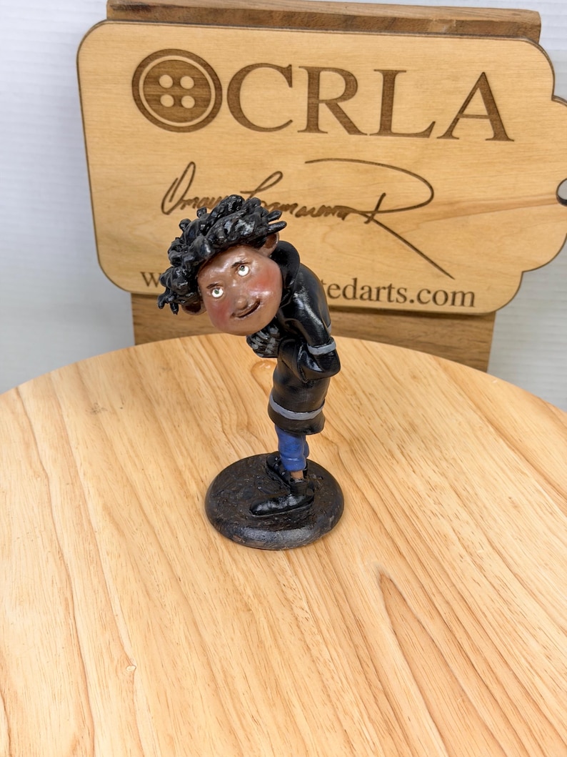 May include: A handcrafted figurine of a person with dark curly hair, wearing a black jacket, blue pants, and black shoes. The figurine is on a black circular base. The background includes a wooden sign with the text "CRLA" and "tedarts.com".