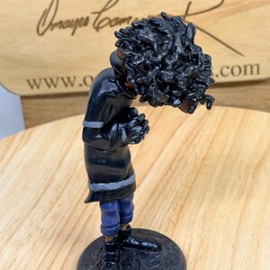 May include: A handcrafted figurine of a person with dark skin, wearing a black coat with gray trim and blue pants. The figure has a large head of black curly hair and is standing on a dark, round base. The figurine is on a wooden surface.