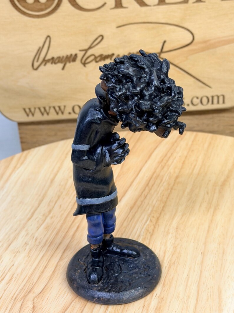 May include: A handcrafted figurine of a person with dark skin, wearing a black coat with gray trim and blue pants. The figure has a large head of black curly hair and is standing on a dark, round base. The figurine is on a wooden surface.