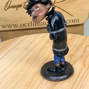 May include: A handcrafted figurine of a person with dark skin, wearing a black coat, blue pants, and black shoes. The figurine has a round base and is posed with a slight lean. The figure has a whimsical, cartoonish style.