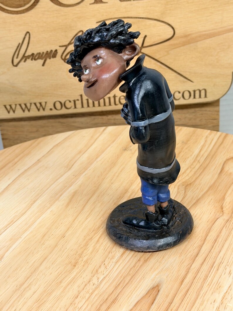 May include: A handcrafted figurine of a person with dark skin, wearing a black coat, blue pants, and black shoes. The figurine has a round base and is posed with a slight lean. The figure has a whimsical, cartoonish style.