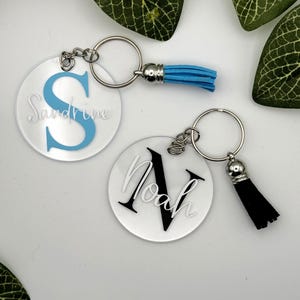 May include: Two circular acrylic keychains with metal rings and tassels. One keychain features a light blue letter S with the name Sandrine in white script. The other keychain features a black letter N with the name Noah in white script.