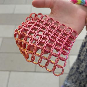 May include: A flexible, rectangular mesh sheet composed of interconnected hexagonal links. The material features a gradient color transition, shifting from a vibrant orange hue on the left side to a bright magenta pink on the right side.
