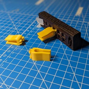 Könnte beinhalten: Ein dunkelbrauner, rechteckiger Holzblock mit einem gitterartigen Muster an der Seite, an dem ein gelber Kunststoffclip befestigt ist, begleitet von zwei weiteren losen gelben Kunststoffclips im gleichen Design und Form.