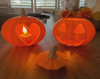 May include: Three orange pumpkin decorations for Halloween. One pumpkin has a heart-shaped cutout with a lit candle inside. Another has a carved face. A small pumpkin stem is in front.
