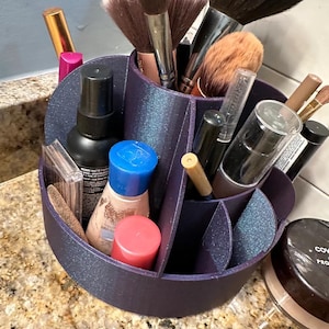 May include: A dark purple makeup organiser with multiple compartments, holding makeup brushes, bottles, and containers. The organiser sits on a speckled countertop, and contains brushes, lipsticks, and powder.