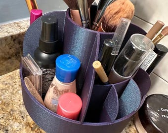 Può includere: Un organizer per trucchi viola scuro a più scomparti pieno di vari pennelli, flaconi e contenitori cosmetici. L'organizer è su un piano di lavoro maculato e contiene pennelli, rossetti e cipria.