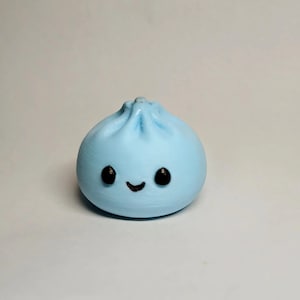 May include: A light blue, rounded dumpling-shaped figurine with a pleated top. The object features two small, black, circular eyes and a thin, black, curved line representing a smiling mouth, all set against a smooth, matte-textured surface.