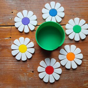Könnte beinhalten: Eine Sammlung von untersetzern in Form von Gänseblümchen mit weißen Blütenblättern und farbigen Zentren in Blau, Grün, Orange, Rot, Gelb und Lila. Ein grüner Becher befindet sich in der Mitte der Untersetzer, auf einer Holzoberfläche.