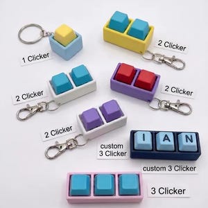 May include: A collection of mechanical keyboard switch keychains featuring one, two, or three keys in various colors including yellow, blue, red, white, and purple. Some include metal lobster clasps and labels reading 1 Clicker, 2 Clicker, 3 Clicker, or custom 3 Clicker with the letters I, A, and N on the keys.