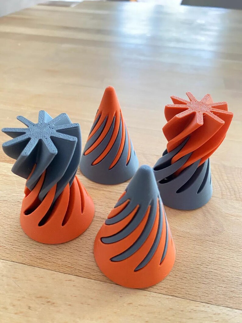 May include: Four abstract, 3D-printed sculptures. Each piece features a cone shape with orange and gray color scheme. The tops of the sculptures have a star-like design. The objects are displayed on a wooden surface.