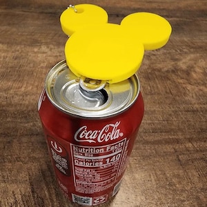 May include: A yellow Mickey Mouse-shaped can opener rests on a red Coca-Cola can. The can opener is made of a solid yellow material and has a small loop for a keyring. The Coca-Cola can displays nutrition information.