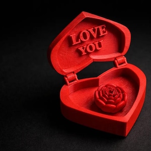Peut inclure: Une boîte rouge en forme de cœur avec les mots "LOVE YOU" sur le couvercle. À l'intérieur de la boîte ouverte se trouve une rose rouge. La boîte est sur une surface noire.