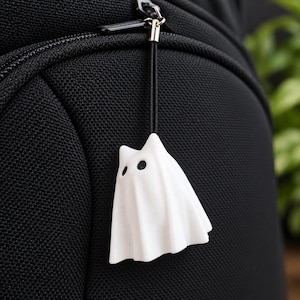 May include: A white ghost-shaped charm with two black circular eyes, attached to a black cord loop. The charm is suspended from a metal ring against a textured black fabric surface, creating a contrast between the white figure and the dark background.
