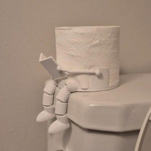 May include: A novelty white toilet roll holder in the shape of a person reading. The holder is made of white plastic and is designed to sit on a toilet. The person has legs and feet hanging over the side, giving a whimsical look.
