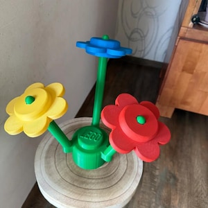 May include: A colorful LEGO flower arrangement featuring three flowers in yellow, red, and blue. The flowers are mounted on green stems and a green base with the LEGO logo. The arrangement sits on a small, round wooden table.