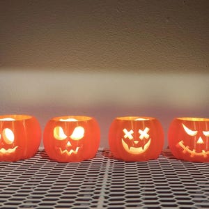 May include: Four orange, illuminated pumpkin-shaped candle holders with carved faces. Each has a unique expression, including a smiling face, a surprised face, and a cross-eyed face. The candle holders are on a metal grate.