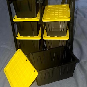 May include: A black storage rack frame holding four black rectangular bins with bright yellow lids. One additional black bin sits in front of the rack with its matching yellow lid placed separately to the side, showing a grid pattern on top.