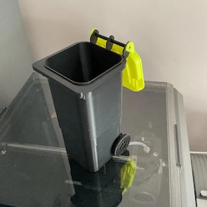 May include: A small, black miniature bin with a hinged lid and wheels, highlighted with bright yellow accents. The small bin is designed to resemble a full-sized waste receptacle. The wheels and lid are yellow, contrasting with the black body.