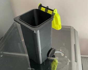 May include: A miniature black trash bin with a hinged lid and wheels, accented with bright yellow details. The small bin is designed to resemble a full-sized trash receptacle. The wheels and lid are yellow, contrasting with the black body.