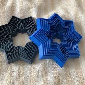 May include: Two star-shaped cookie cutters, one dark gray and one bright blue, are nested together. Each cutter has multiple layers, creating a three-dimensional effect. The cutters are made of plastic and sit on a textured, off-white surface.