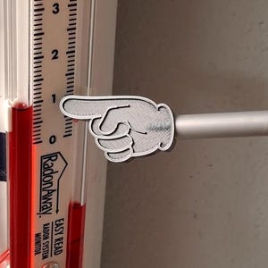 May include: A gray, 3D-printed hand pointing left, attached to a silver rod. The hand is positioned near a RadonAway radon monitor, with a red liquid level indicator and a scale marked from 0 to 3.