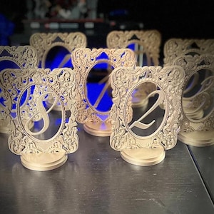 May include: A collection of gold-coloured table number stands featuring intricate, ornate, laser-cut rectangular frames with oval centres. Each stand displays a large, stylised numeral, including the numbers eight and two, mounted on a circular base.