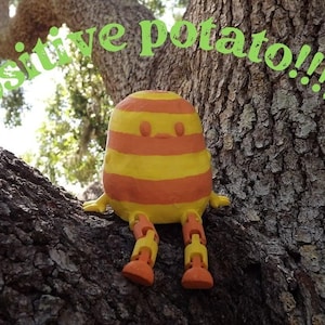 May include: A whimsical, handmade sculpture of a potato-like character with yellow and orange stripes, perched on a tree branch. The text "positive potato!!!!" is arched above the figure. The figure has articulated legs.