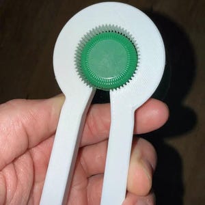 May include: A white bottle opener with a green bottle cap held in place. The opener has a circular design with gear-like teeth and two long handles. The bottle cap is green and has a ridged design.