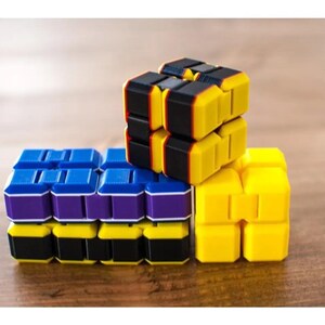 May include: A close-up of three colorful fidget cubes. The cubes are yellow, black, blue, and purple. The cubes are stacked on top of each other on a wooden surface.