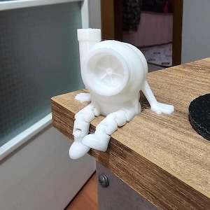Könnte beinhalten: Eine weiße, 3D-gedruckte Figur eines Turboladers mit Beinen und Armen, die auf einer Holzoberfläche sitzt. Die Figur hat ein Turbinenrad im Gehäuse und ruht auf einer braunen Holzoberfläche.