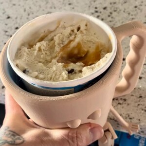 Puede incluir: Un soporte beige para helado impreso en 3D con un asa, dise&ntilde;ado para sostener una pinta de helado. El soporte contiene una pinta de helado de color marr&oacute;n claro y chispas de chocolate. El soporte tiene un dise&ntilde;o ergon&oacute;mico &uacute;nico.