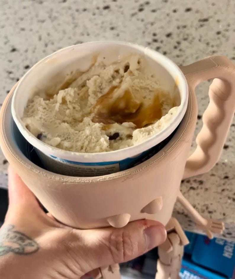 Puede incluir: Un soporte beige para helado impreso en 3D con un asa, dise&ntilde;ado para sostener una pinta de helado. El soporte contiene una pinta de helado de color marr&oacute;n claro y chispas de chocolate. El soporte tiene un dise&ntilde;o ergon&oacute;mico &uacute;nico.