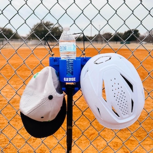 May include: A blue baseball equipment holder attached to a chain-link fence, holding a water bottle, a white baseball cap with a black brim, a baseball bat, and a white baseball helmet. The word "GAUGE" is printed on the holder.