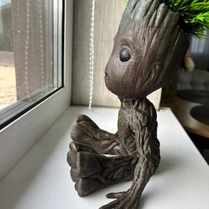 以下が含まれることがあります： 茶色と緑色の3Dプリントされたグルートのプランターに、小さな鉢植えの植物が入っています。プランターは、ガーディアンズ・オブ・ギャラクシー映画のキャラクター、グルートの形をしています。プランターは白い表面の上に置かれています。
