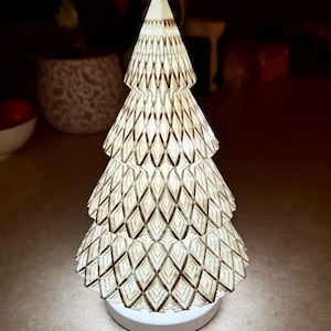 May include: An illuminated Christmas tree ornament. The tree is constructed from layered paper with a diamond pattern, giving off a warm light. It stands on a white, circular base. The design is intricate and detailed, ideal for festive home decoration during the holiday season.