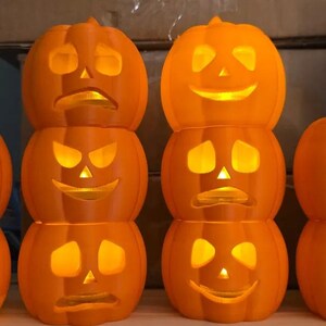 May include: Orange Halloween pumpkin decorations with carved faces and internal lights. Each pumpkin has a unique expression, stacked in pairs. The illuminated faces create a festive and spooky ambiance, perfect for seasonal decor.