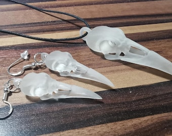 May include: A set of frosted glass bird skull jewelry, including a necklace and earrings. The necklace has a black cord and the earrings have silver hooks.