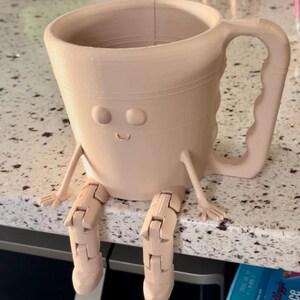 Puede incluir: Una taza de caf&eacute; beige impresa en 3D con una cara sonriente, brazos y piernas articuladas. La taza tiene un asa grande y est&aacute; sobre una encimera. La taza es un art&iacute;culo decorativo caprichoso y &uacute;nico.