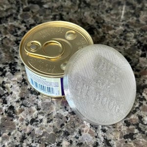 Peut inclure: Une boîte de conserve alimentaire en métal doré avec un couvercle à anneau et un couvercle en plastique translucide texturé, de forme circulaire, portant le texte en relief "PET FOOD" sur sa surface.