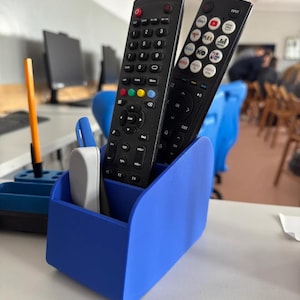May include: A blue desk tidy containing two black remote controls and a white pen. The organiser has three compartments and is on a white surface. The remotes have multiple buttons and are angled towards the viewer.