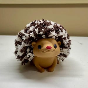 May include: A charming hedgehog ornament with a brown body and a pom-pom-style back in brown and white. The hedgehog has blue eyes and a red nose, creating a handcrafted aesthetic. Suitable for home decor, this ornament is a delightful addition.