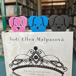 May include: Three dog-shaped bookmarks in pink, blue, and gray are placed on top of a book. The book's title is "Jodi Ellen Malpasová" and the cover has decorative elements. The bookmarks are made of a textured material.