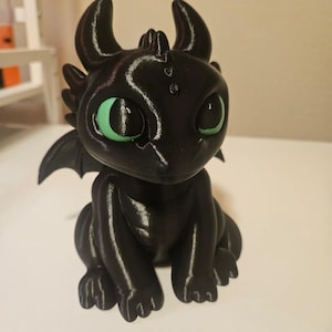 以下が含まれることがあります： 大きな緑色の目、小さな翼、角を持つ黒いドラゴンの置物。 ドラゴンは足を体の下に折りたたんで座っています。 置物は光沢のある滑らかな素材で作られています。